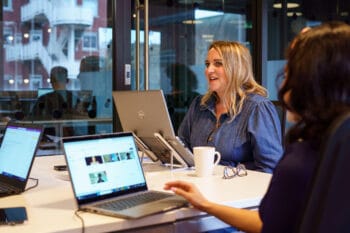 A colleague sat at a desk speaking to another colleague who is off camera to the right. Another female colleague is sitting on the left in front of a laptop