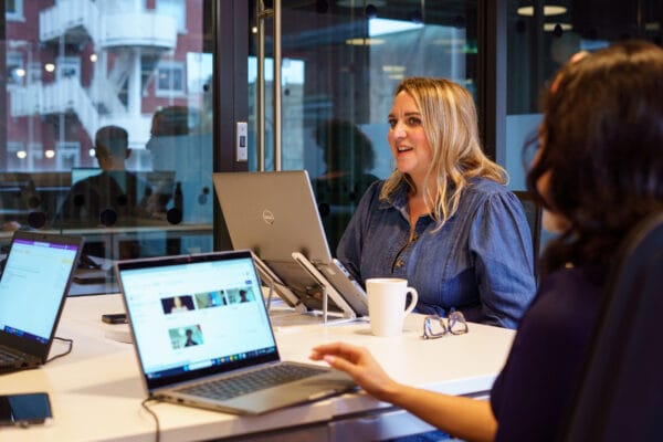 A colleague sat at a desk speaking to another colleague who is off camera to the right. Another female colleague is sitting on the left in front of a laptop