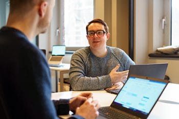 two men talking at a table with a laptop